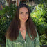 A woman with long brown hair and a green patterned shirt stands outside in front of lush, green tropical plants, smiling softly at the camera.