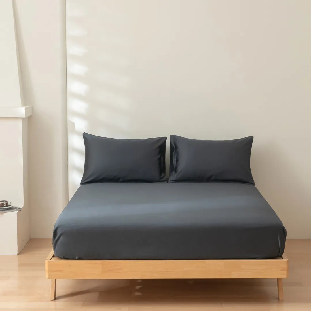 A neatly made bed with a Linenly Luxe Sateen Fitted Sheet in Charcoal on a simple wooden frame, positioned in a room with soft natural lighting casting gentle shadows on a light-colored wall.