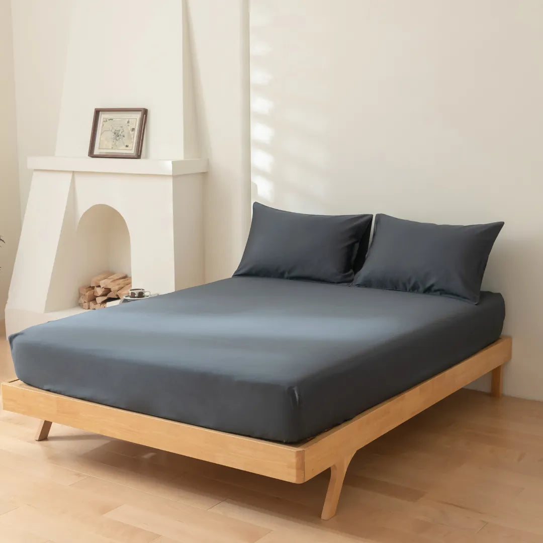 A neatly made bed with a Linenly Luxe Sateen Fitted Sheet in Charcoal in a minimalist bedroom, featuring a decorative faux fireplace with logs and a framed picture on the mantel.