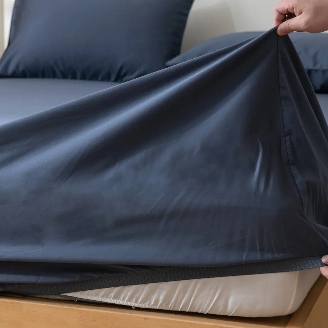 A hand pulls a Linenly Luxe Cotton Sateen Fitted Sheet in Midnight over a mattress on a wooden bed, with matching pillowcases and luxury bedding featured in the background.