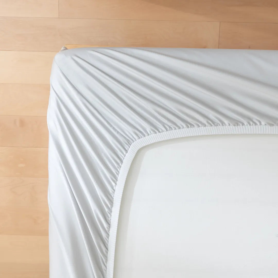A neatly made bed with a Linenly Luxe Sateen Fitted Sheet in Silver on a wooden floor, exhibiting a minimalistic and tidy bedroom setting.
