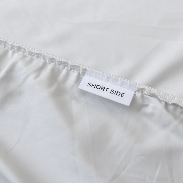 A close-up of a Linenly Luxe Sateen Fitted Sheet in Silver, made of long-staple cotton, with a tag labeled "short side" to indicate the orientation of the sheet for easier bed-making and a luxurious sleep experience.
