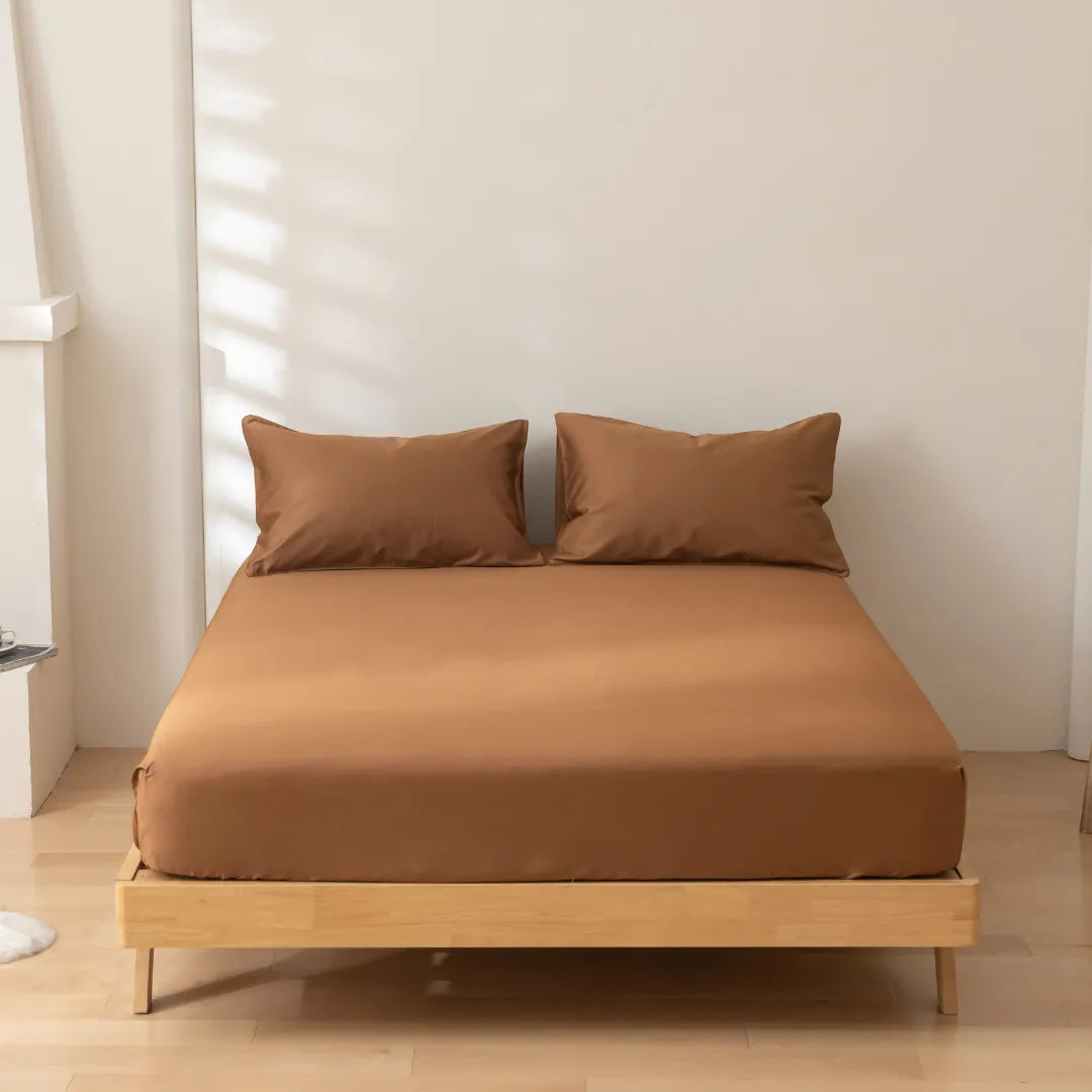 A neatly made bed with a wooden frame and Linenly Luxe Sateen Fitted Sheet in Terracotta in a room with soft natural light casting shadows on the wall.