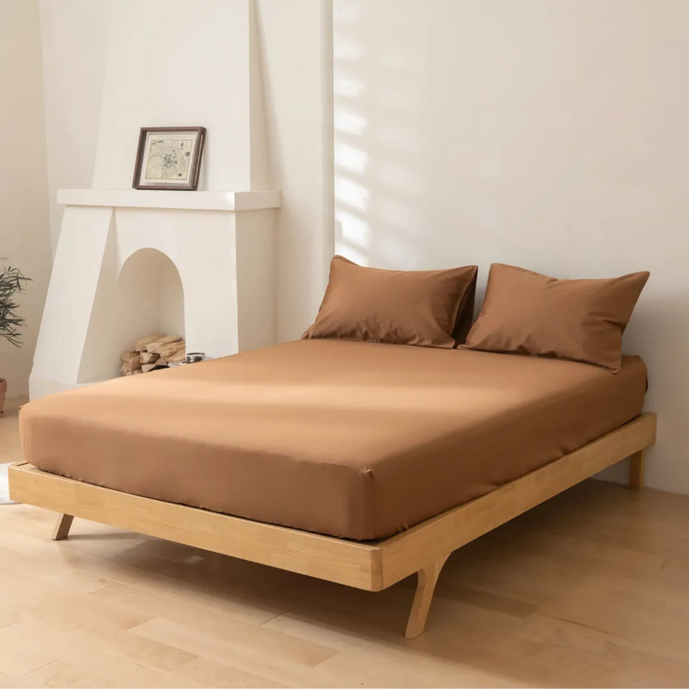 A minimalist bedroom with a low-profile wooden bed frame and matching earth-toned bedding, complemented by a light and airy interior with a white fireplace and frame decor. The addition of a Linenly Luxe Sateen Fitted Sheet in Terracotta.