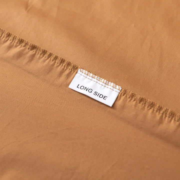 A close-up view of a Linenly Luxe Sateen Terracotta Fitted Sheet with a tag labeled "long side" indicating the orientation of the long-staple cotton material.