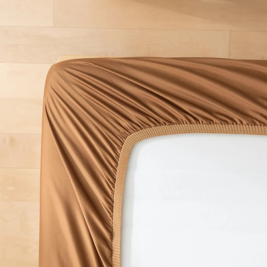 A neatly draped Luxe Sateen Fitted Sheet in Terracotta by Linenly on a corner of a mattress, highlighting a luxurious and smooth texture atop a wooden floor, complements the cozy bedroom ambiance.