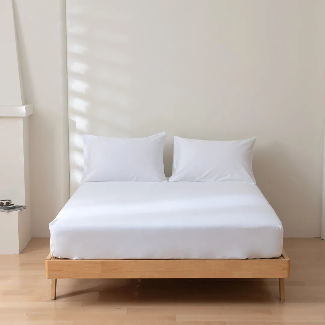 A neatly made bed with a Linenly Luxe Sateen Fitted Sheet in White on a simple wooden bed frame in a bright minimalist room.