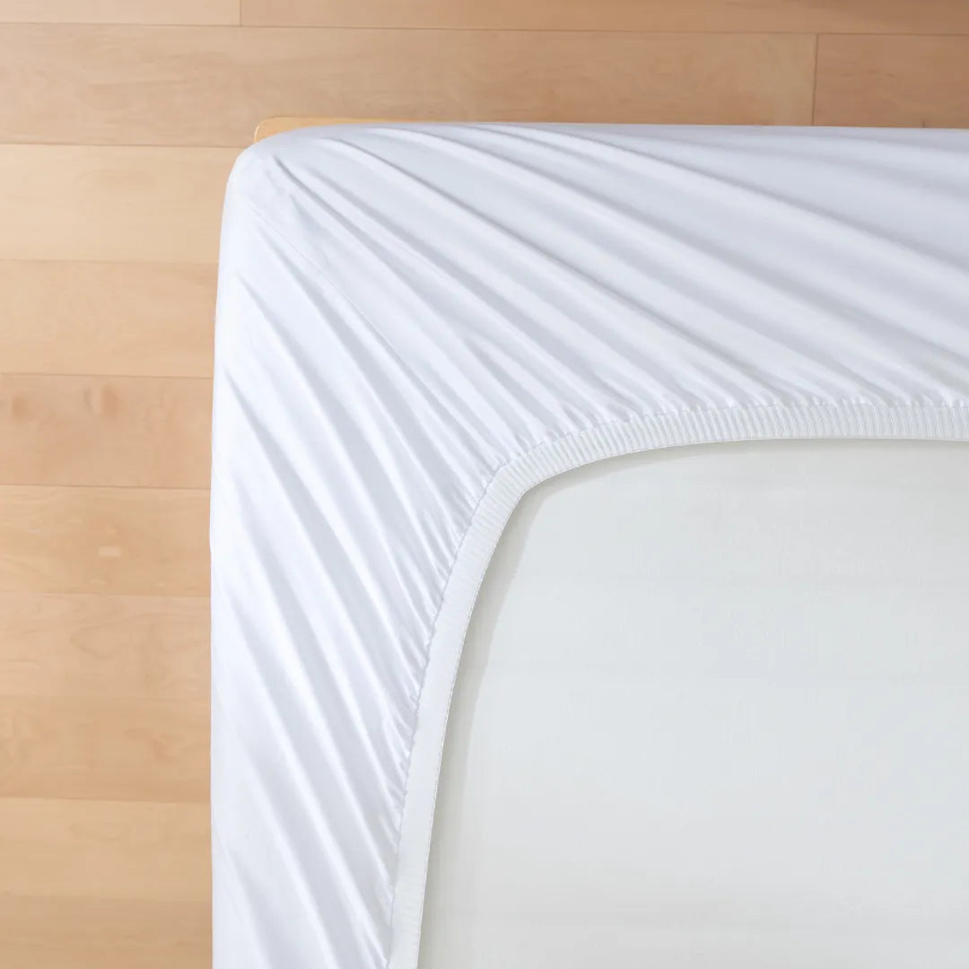 A neatly fitted Linenly Luxe Sateen Fitted Sheet - White on a bed with a wooden headboard, showcasing a clean and simplistic bedroom aesthetic.