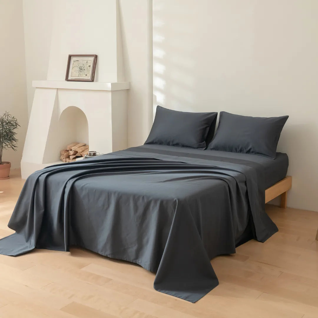 Modern minimalist bedroom with a neatly made bed featuring a Linenly Luxe Sateen Flat Sheet in Charcoal, made of premium long staple cotton, in a serene space with a white fireplace and framed art.