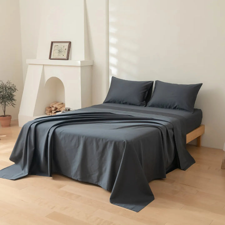 A neatly made bed with a Linenly Luxe Sateen Flat Sheet in Charcoal, made of premium long staple cotton in a sateen weave, positioned in a minimalist bedroom with light walls, wood flooring, a small plant, and a decorative.