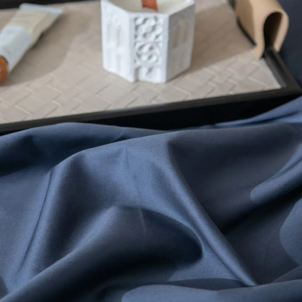A serene composition featuring Linenly's Luxe Sateen Flat Sheet in Midnight, a premium long staple cotton, gracefully draped over an edge, with a white textured cup and a partially seen book resting quietly in the background, creating an ambiance.