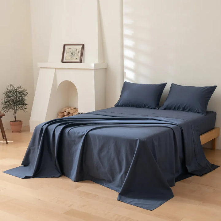 A neatly made bed with a Linenly Luxe Sateen Flat Sheet in Midnight, in 500 thread count premium long staple cotton, in a serene bedroom with minimalist decor, featuring a faux fireplace and a potted plant, bathed in