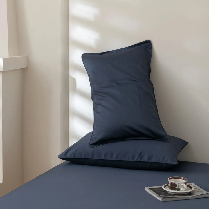 A tranquil corner featuring a navy blue pillow leaning against the wall on a Linenly Luxe Sateen Flat Sheet in Midnight, with a cup of coffee and reading material on the side, bathed in the soft light filtering through blinds.