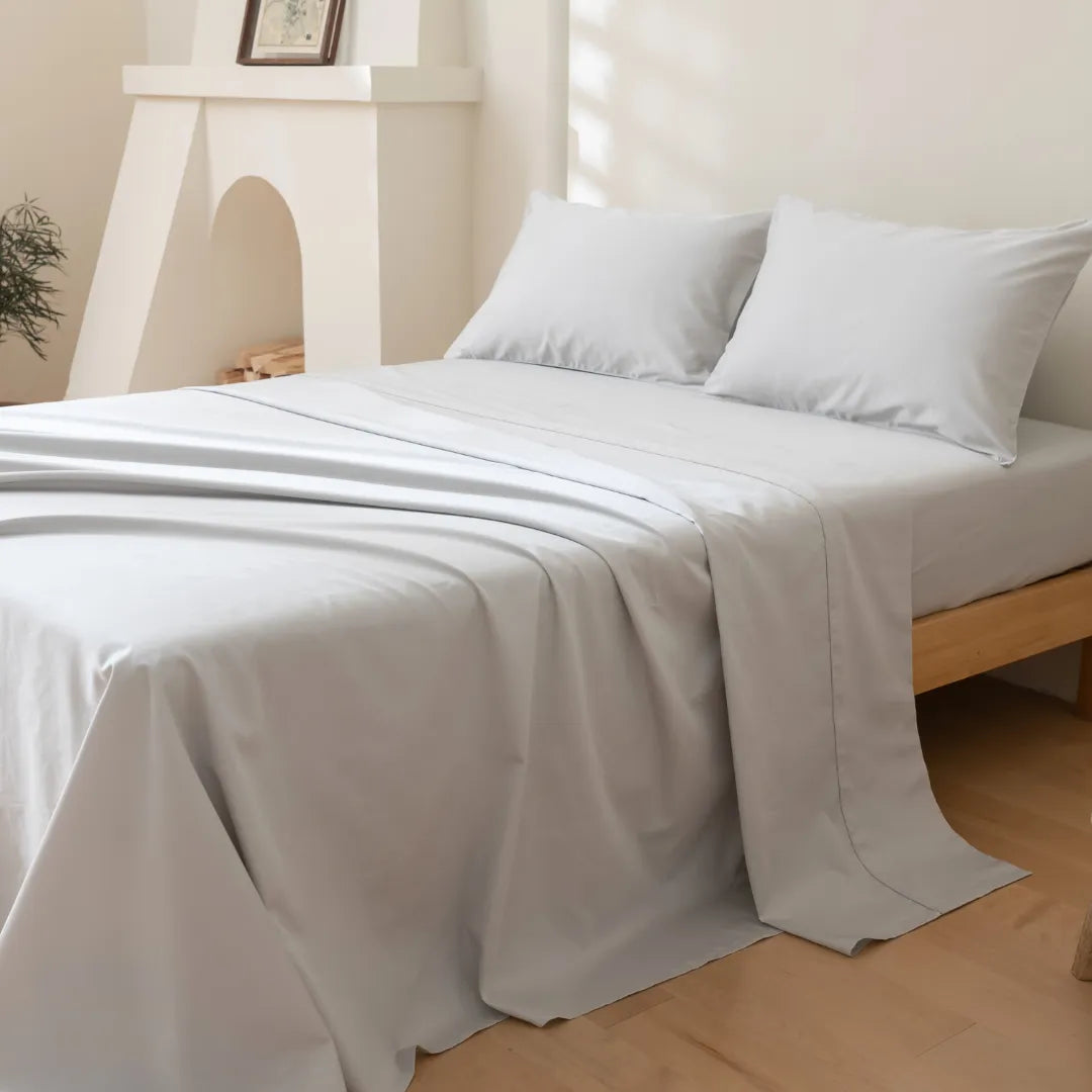 A neatly made bed features the Linenly Luxe Cotton Sateen Flat Sheet in Silver and two pillows on a wooden frame. Light walls, floors, a small plant, and framed picture complete the bright, minimalistic room’s serene atmosphere.