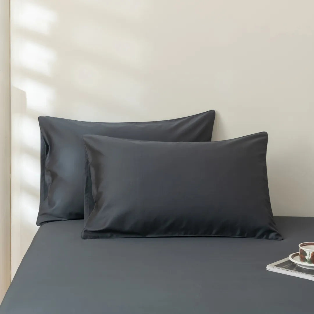 Two Linenly Luxe Cotton Sateen Pillowcases in Charcoal rest against a matching sheet, with sunlight casting soft shadows on the neutral wall. A small tray with a cup and saucer sits at the bed's edge.