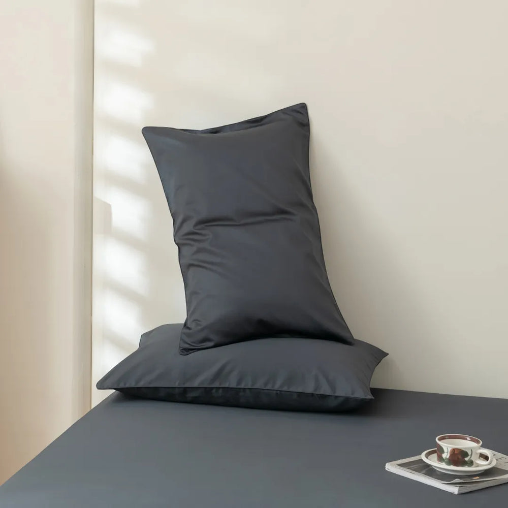 A minimalist setting with a neatly arranged dark pillow, enveloped in Linenly's Luxe Sateen Pillowcase Set in Charcoal, on top of another against a pale wall, beside a small tray with a cup and saucer.