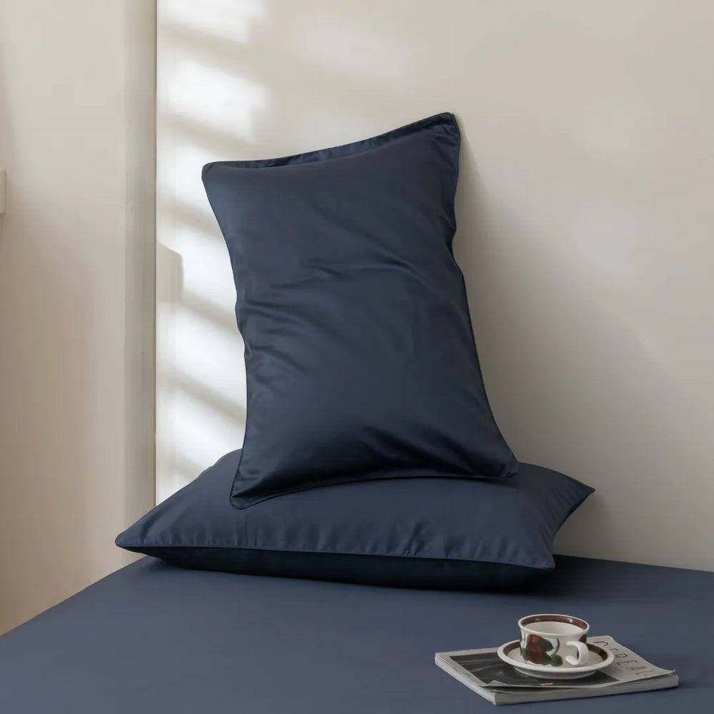 Minimalist interior with a neatly placed Linenly Luxe Sateen Pillowcase Set - Midnight featuring an envelope closure design against a softly lit wall, accompanied by a matching cushion on a sleek surface with a cup of coffee and reading materials, inviting