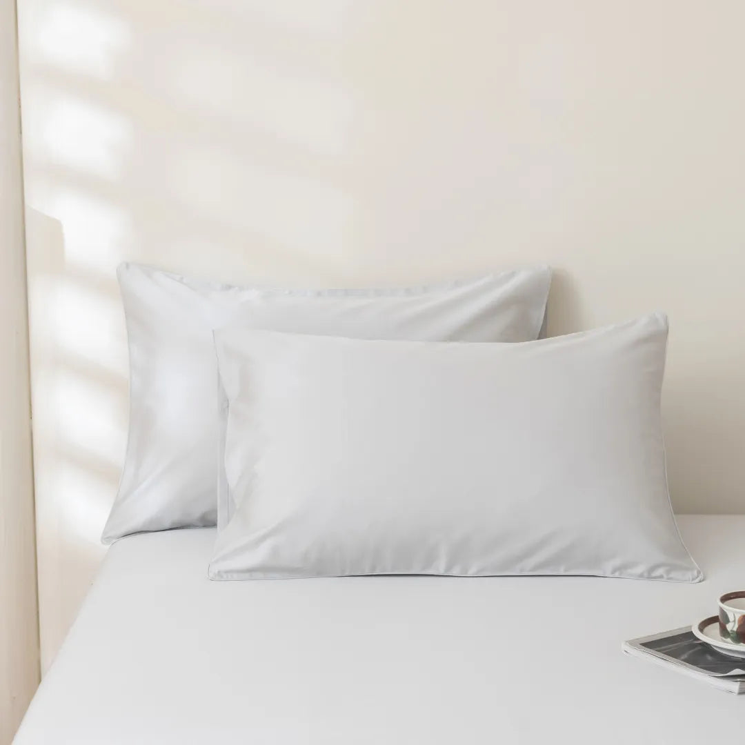 Two white pillows encased in Linenly Luxe Sateen Pillowcase Set - Silver rest on a neatly made bed with sunlight softly filtering through blinds.