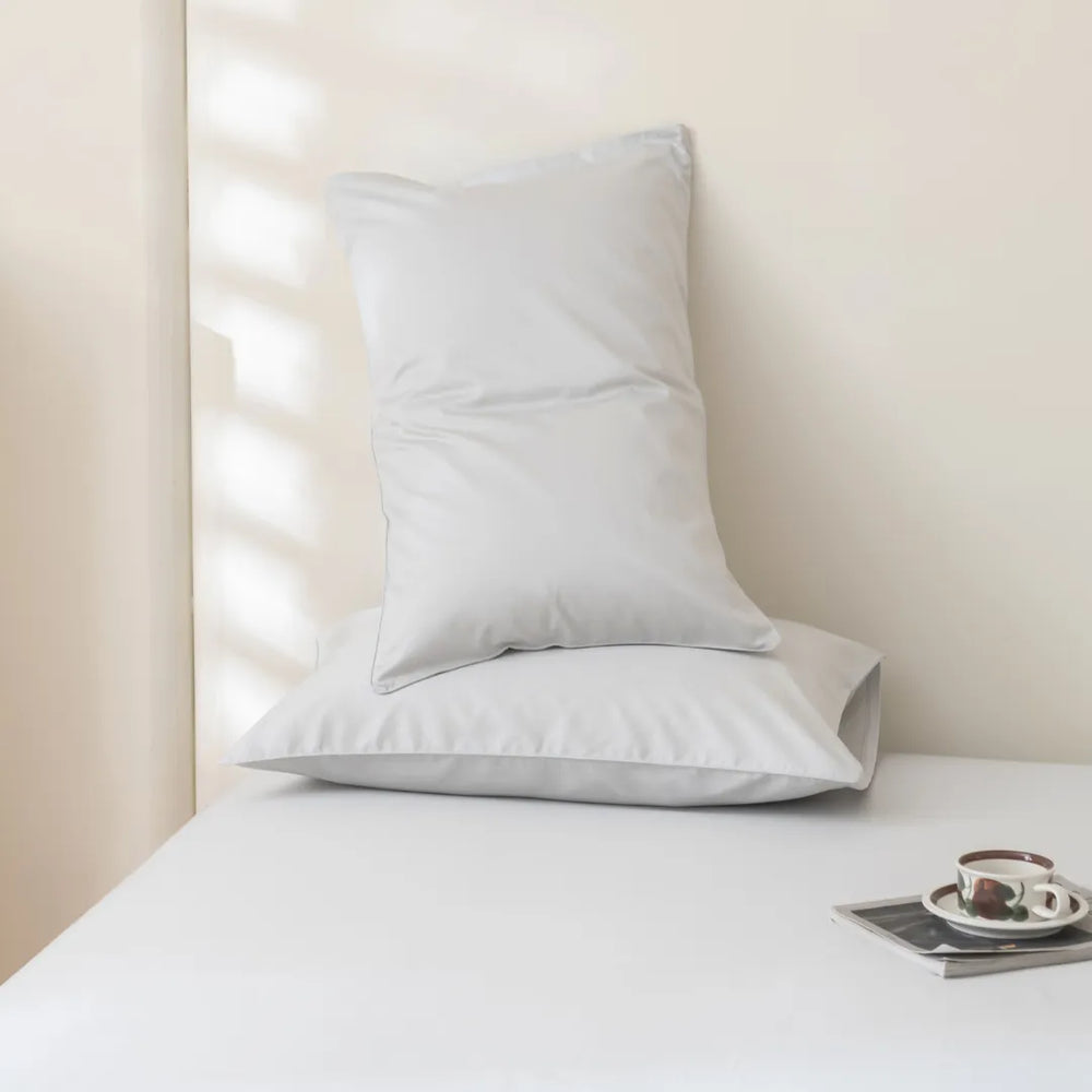 A serene corner with a neatly arranged Luxe Sateen Pillowcase Set - Silver by Linenly on a white surface, accompanied by a magazine and a cup of coffee on a small tray, beside a window with soft light.