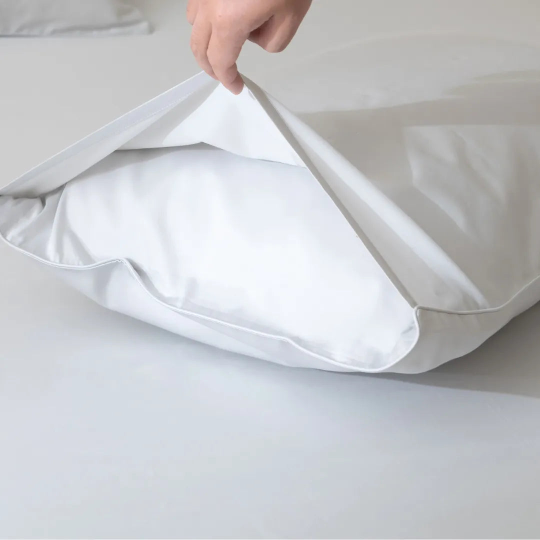 A hand opens the zipper of a Linenly Luxe Cotton Sateen Pillowcase Set in Silver, revealing a pillow inside, all arranged on a pristine white surface.