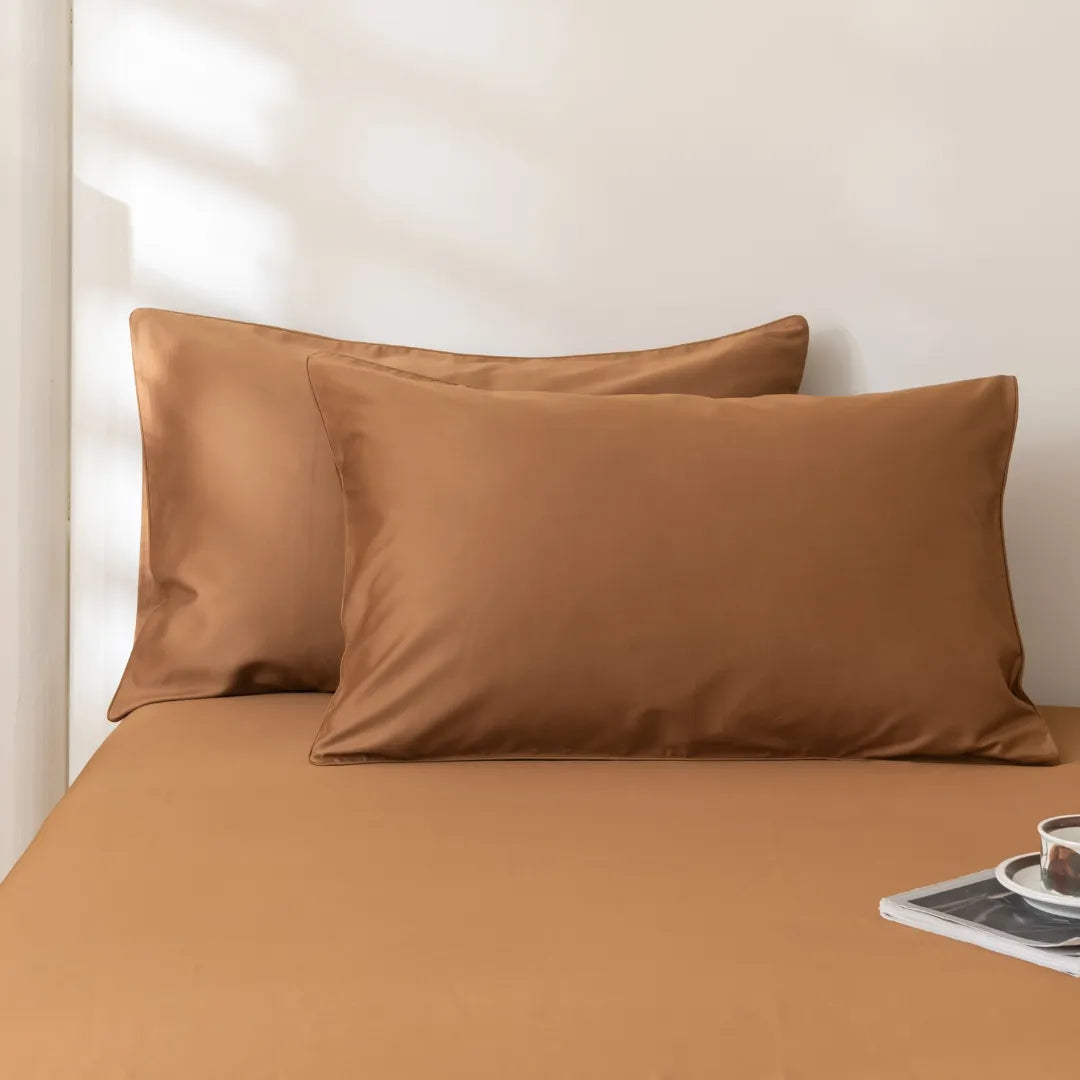 Two smooth, caramel-colored Linenly Luxe Sateen Pillowcase Sets in Terracotta on a neatly made bed with premium bedding, with soft sunlight filtering in, creating a tranquil and cozy atmosphere. A magazine and a cup of coffee on
