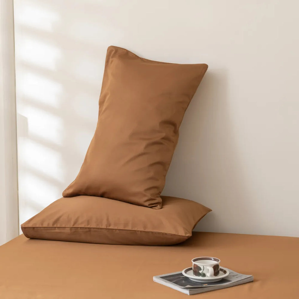 A cozy corner with a Linenly Luxe Sateen Pillowcase Set in Terracotta propped against a wall on a tan surface next to a small stack of magazines with a cup of coffee on top, bathed in soft light.