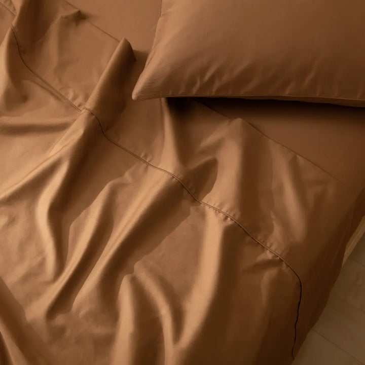 A neatly made bed with smooth, terracotta-colored, long staple cotton Luxe Sateen pillowcases from Linenly bathed in soft, warm light creating gentle shadows on the fabric.