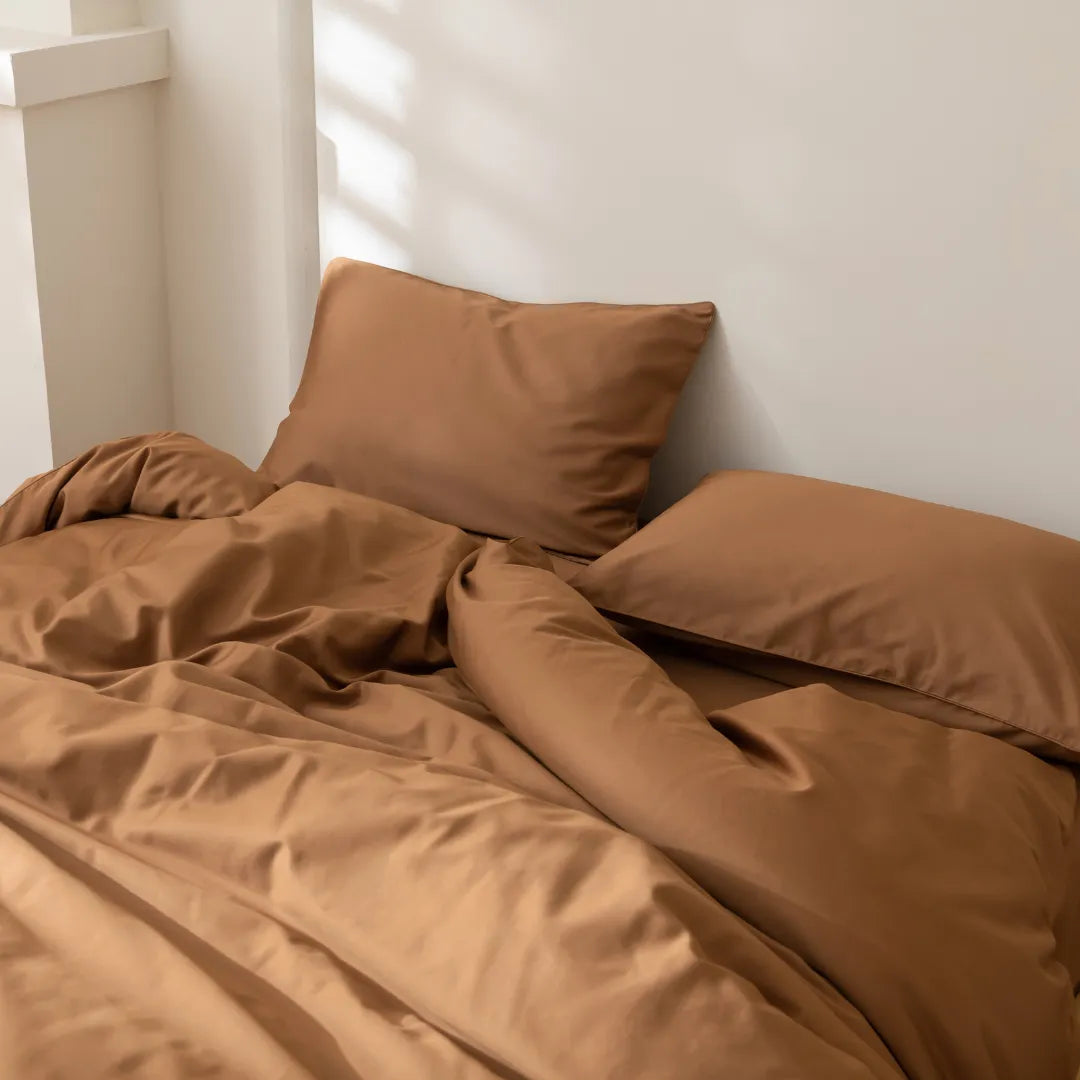 A neatly made bed with Linenly's Luxe Sateen Pillowcase Set in Terracotta basked in soft natural light, creating a cozy atmosphere.