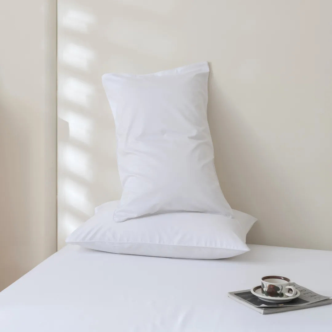 A pristine white Luxe Sateen Pillowcase Set from Linenly with an envelope closure design and cushion set against a pale wall with soft light casting linear shadows, accompanied by a neatly placed coffee cup and saucer on a serene tabletop.