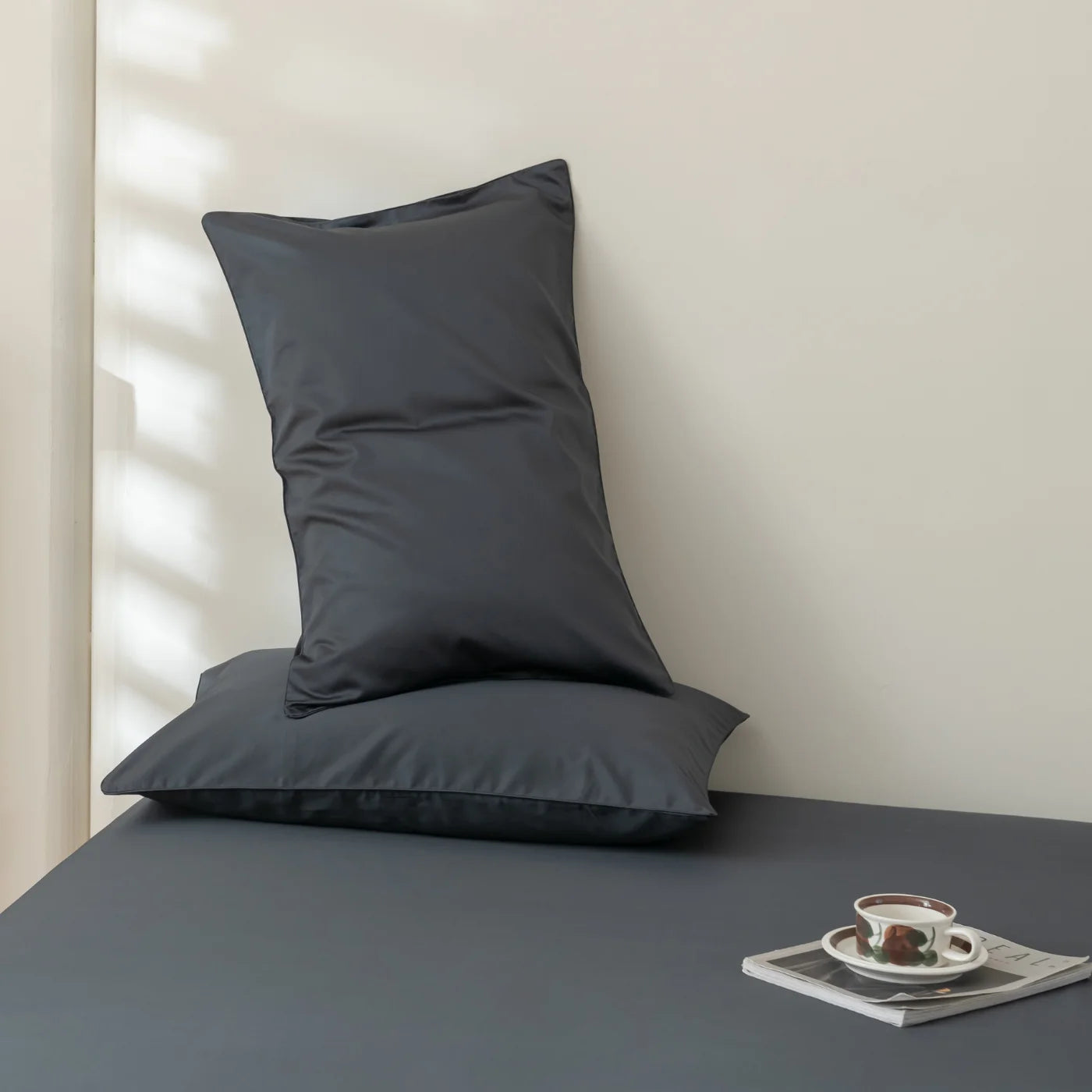 Two dark gray pillows are arranged on a matching dark gray surface against a light-colored wall. A cup of coffee and a saucer sit on a stack of magazines nearby. Soft light and shadows fall across the scene.