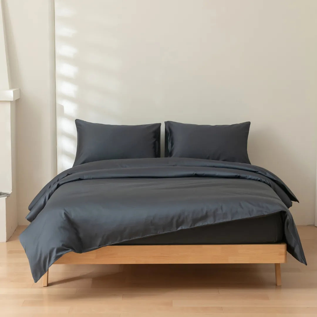 A neatly made bed features Linenly’s Luxe Cotton Sateen Quilt Cover in Charcoal with matching pillows on a light wood frame, set in a minimalist, sunlit room with light wood flooring and an off-white wall.