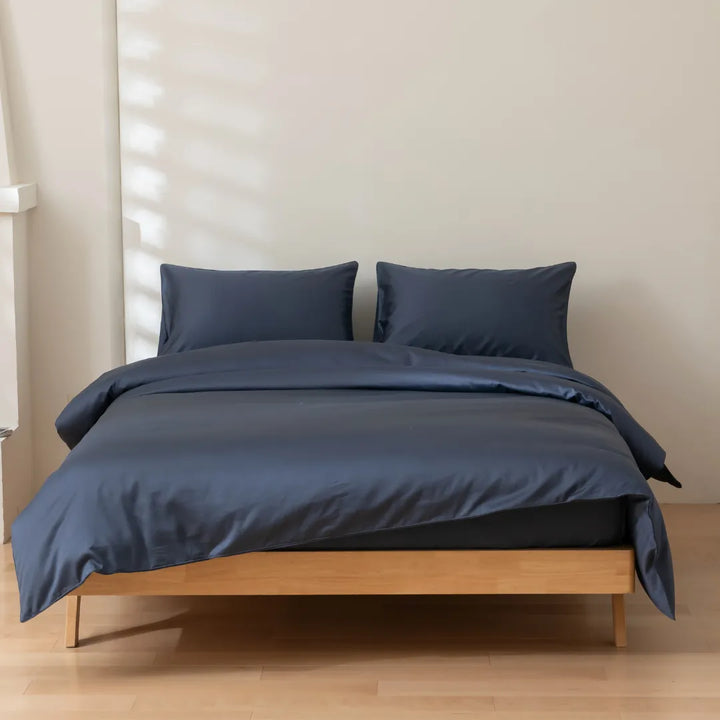 A neatly made bed with a Luxe Sateen Quilt Cover in Midnight from Linenly and two matching pillows on a simple wooden bed frame in a room with soft natural lighting.