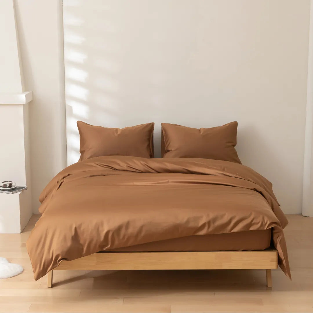 A neatly made bed with a smooth brown bedding set in a minimalist bedroom features light walls and morning light casting soft shadows. The Linenly Luxe Sateen Quilt Cover in Terracotta has a sateen weave, adding an elegant touch.
