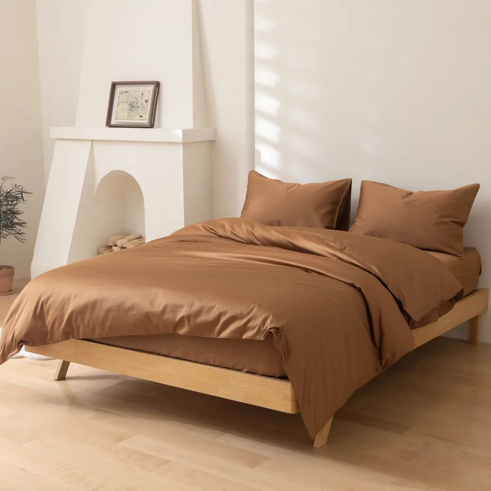 A neatly made bed with a smooth brown Linenly Luxe Sateen Quilt Cover - Terracotta 500 thread count, sateen weave bedsheet set in a minimalist bedroom with soft natural light filtering through the blinds, a simple wooden bed frame.
