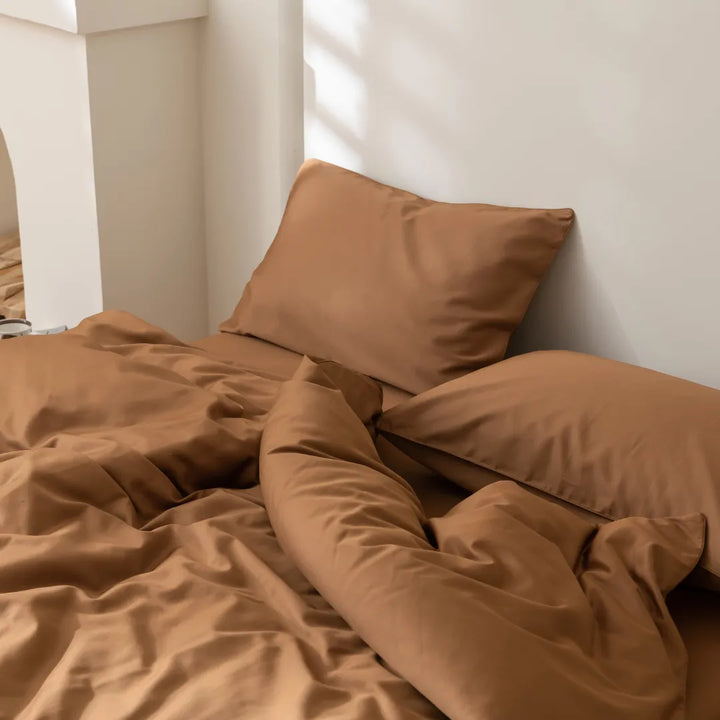 A cozy, unmade bed bathed in soft light, featuring crumpled caramel-colored sheets and a Linenly Luxe Sateen Quilt Cover in terracotta, inviting a moment of relaxation.