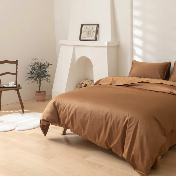A cozy bedroom corner featuring a neatly-made bed with earth-toned linens, including a Luxe Sateen Quilt Cover in Terracotta of 500 thread count in a sateen weave, a classic wooden chair from Linenly.