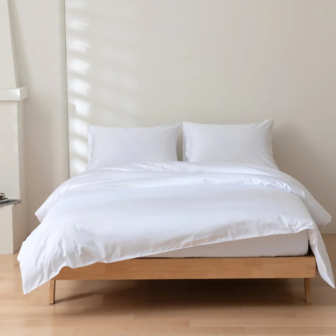 A neatly made bed with a wooden frame and Linenly Luxe Sateen Quilt Cover in White in a bright minimalist room.