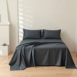 A neatly made bed features the Luxe Cotton Sateen Sheet Set in Charcoal by Linenly and two matching pillows, set against a plain white wall in a minimalist room with light wood flooring and soft window-lit shadows.