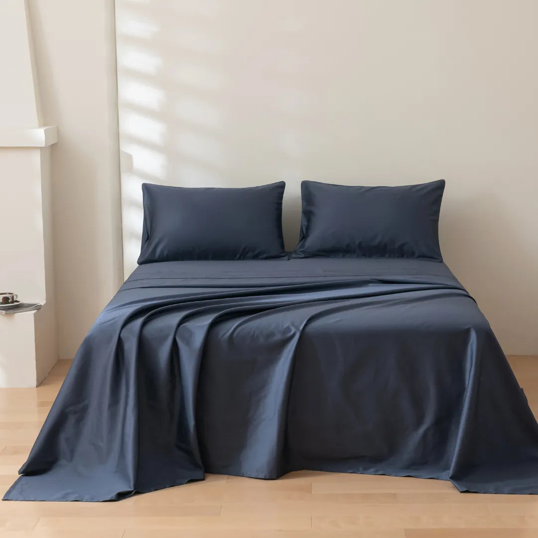 A neatly made bed with Linenly's Luxe Sateen Sheet Set in Midnight, featuring dark blue, high thread count bedding in a serene beige bedroom, bathed in natural light through the window blinds.