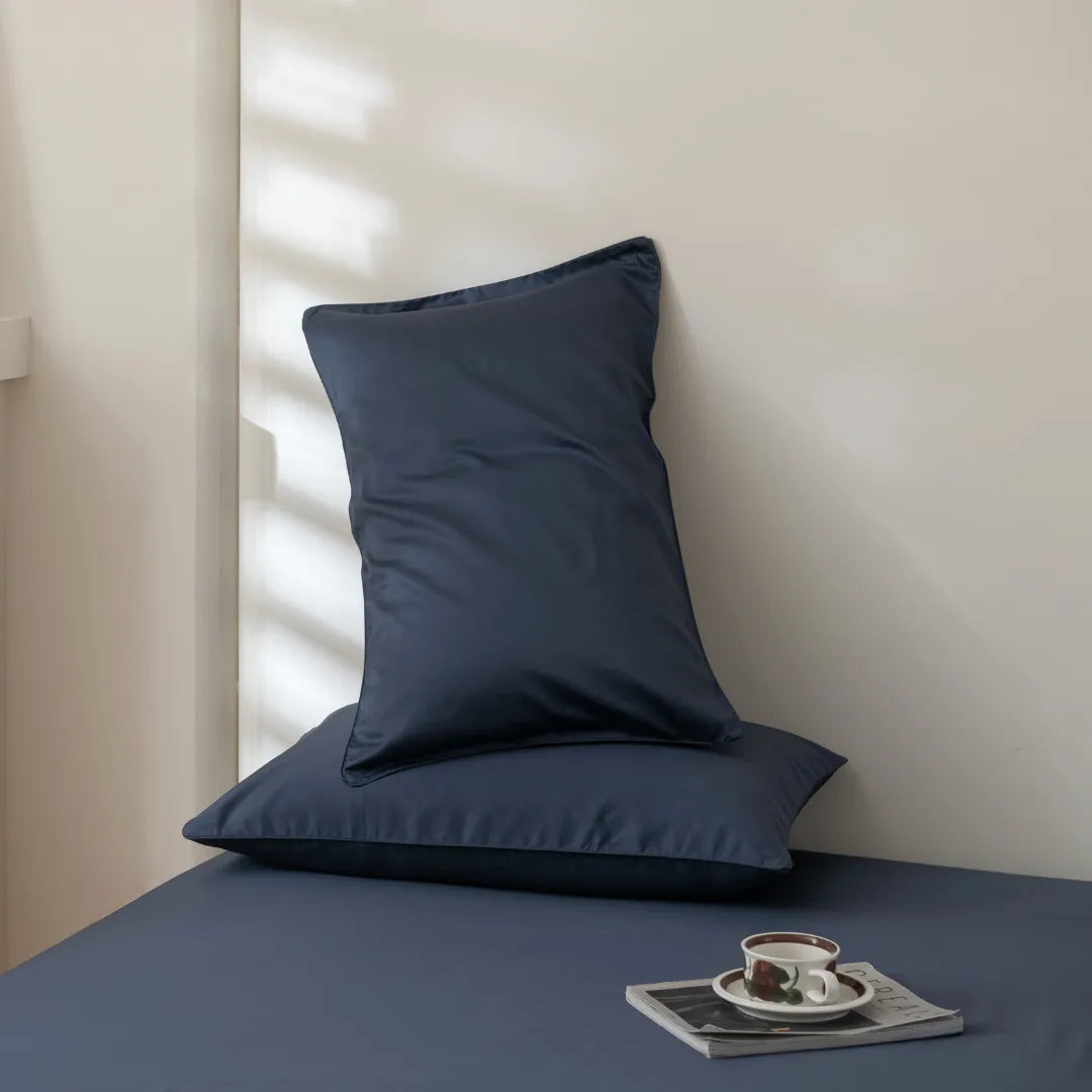 Two navy blue pillows rest against a cream wall on a matching blue bed dressed in the Linenly Luxe Cotton Sateen Sheet Set in Midnight. Sunlight streams in, casting gentle shadows as a cup of coffee sits on a magazine nearby.
