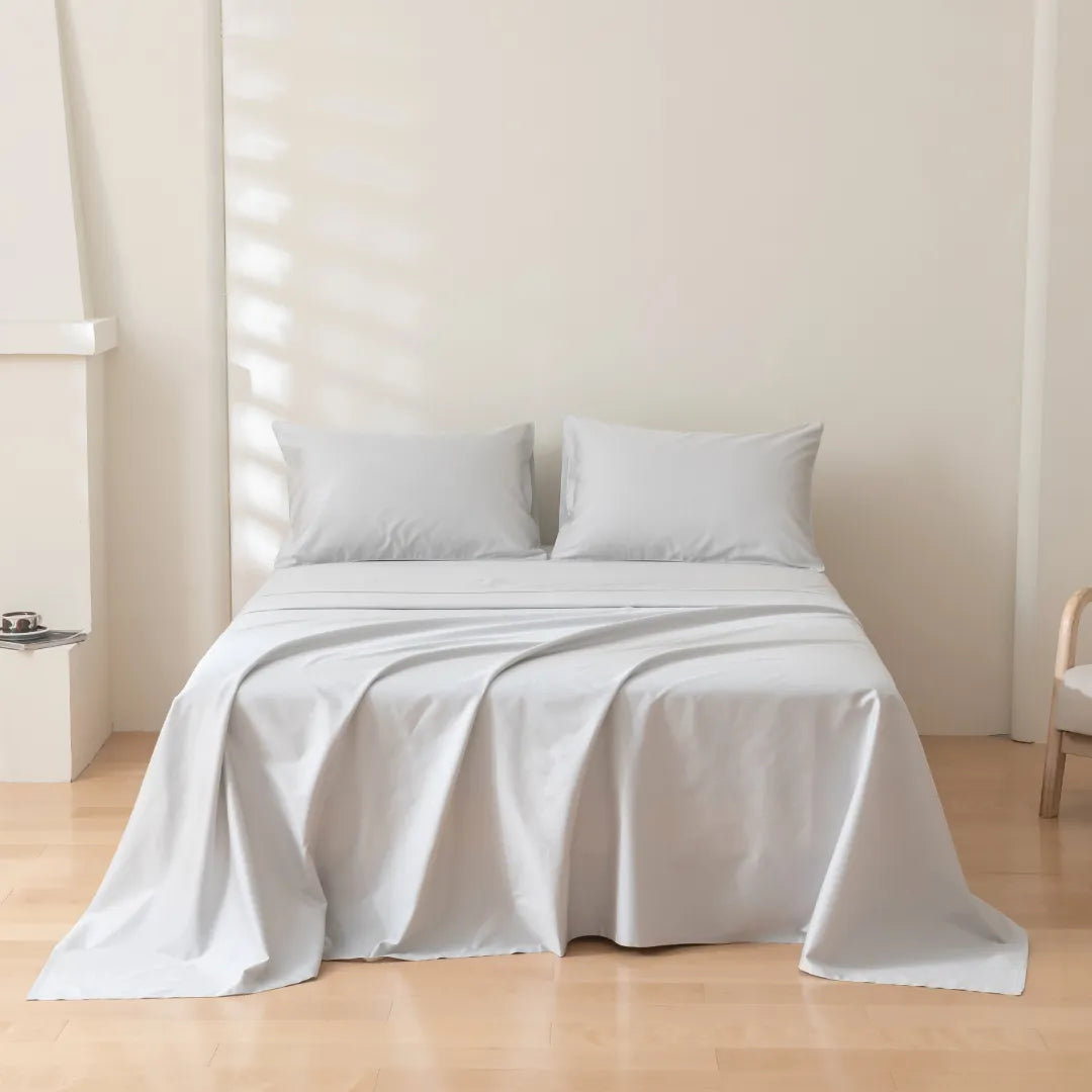A neatly made bed with a smooth light gray duvet and two matching pillows in a bright, serene room with soft natural light filtering through the window, complemented by a Linenly Luxe Sateen Sheet Set - Silver.