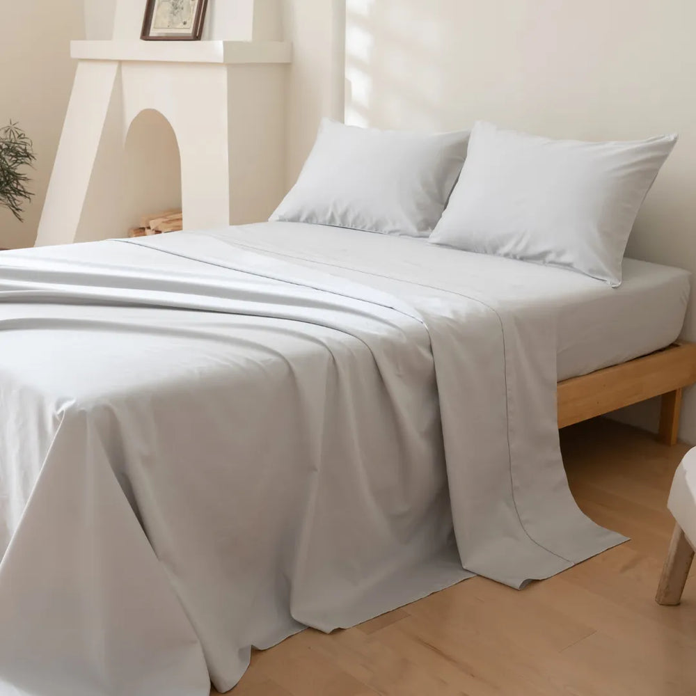 A neatly made bed with a Linenly Luxe Sateen Sheet Set in Silver and two pillows in a bright, serene bedroom with wooden furniture and a minimalist decor.