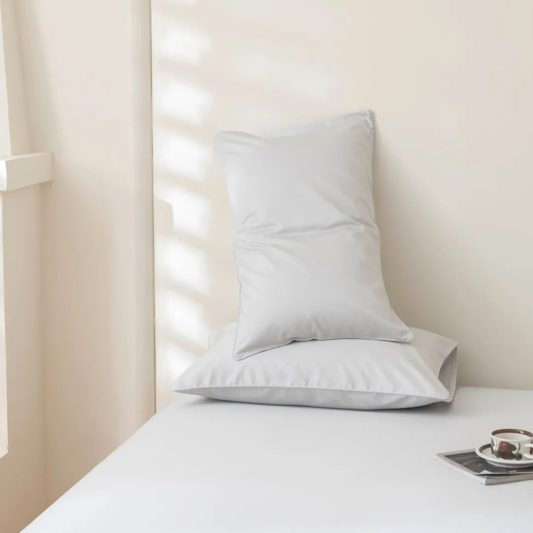 Two white pillows rest on a neatly made bed featuring the Luxe Cotton Sateen Sheet Set in Silver by Linenly. A cup and saucer on a tray complete the tranquil setting as soft light filters through the window.