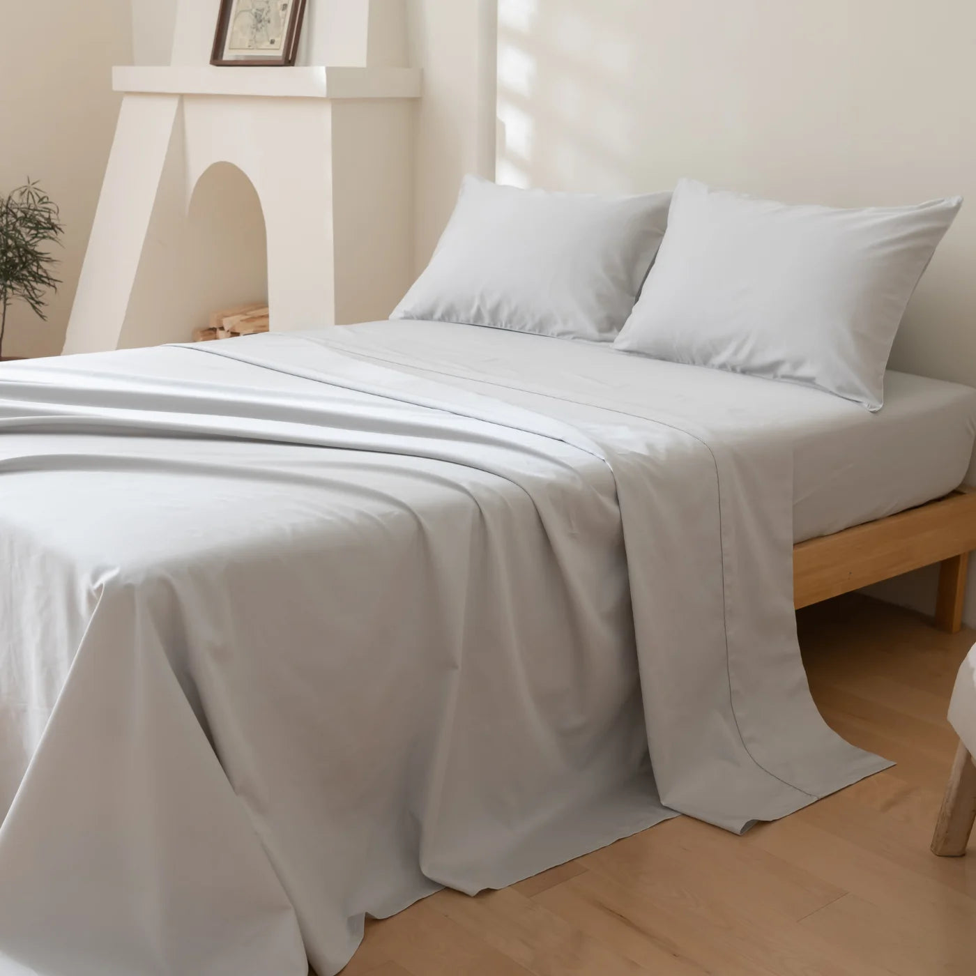 A neatly made bed with light gray sheets and two pillows sits on a wooden frame in a bright, minimalist bedroom with a white fireplace and a small plant in the corner.