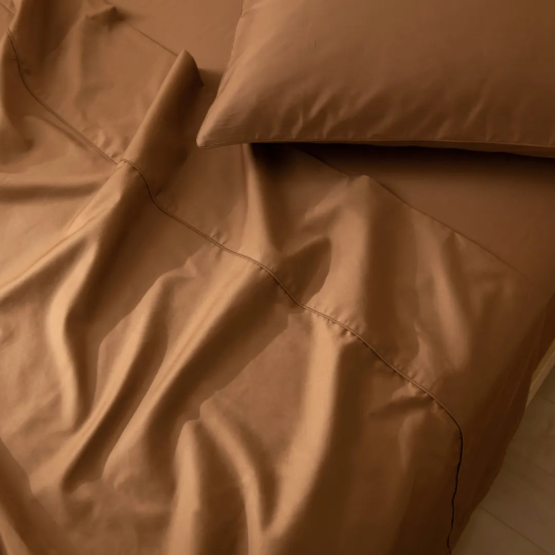 A close-up view of a rumpled Linenly Luxe Sateen Sheet Set in Terracotta on a bed, bathed in soft, warm light, evoking a cozy and inviting atmosphere.