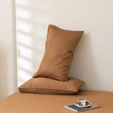Two brown pillows rest on a matching bed made up with Linenly's Luxe Cotton Sateen Sheet Set in Terracotta; one pillow lies flat, the other upright against the wall. A cup and saucer sit on a magazine nearby as soft sunlight casts gentle shadows.