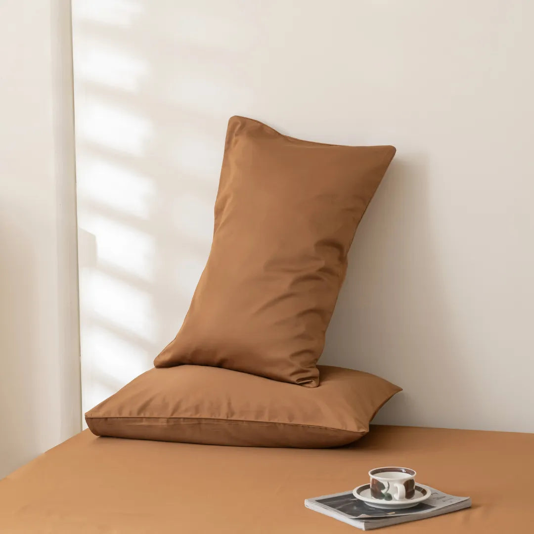 A serene corner featuring a plush burnt orange pillow propped against a soft-toned wall, accompanied by a neatly placed coffee cup on a saucer atop a simple book, all resting on Linenly's Luxe Sateen Sheet Set in Terracotta.