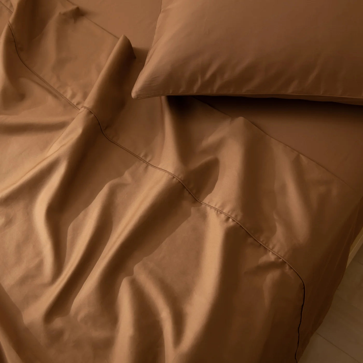 A close-up of a bed with wrinkled brown sheets and a matching brown pillow, showing smooth fabric and soft folds.