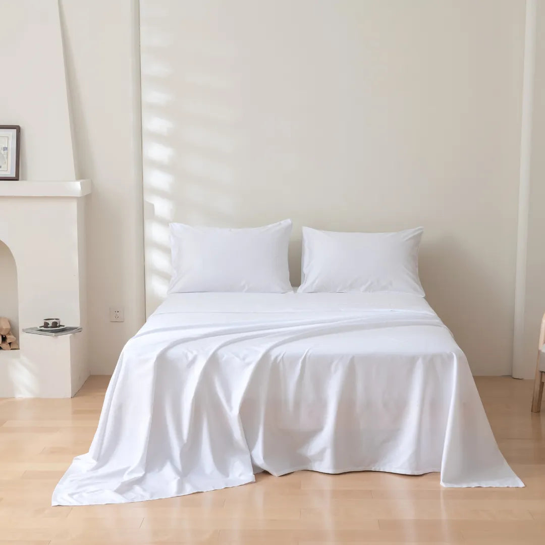 A neatly made bed with two white pillows and the Linenly Luxe Cotton Sateen Sheet Set in a minimalist room with light walls and wooden floor. Sunlight casts soft shadows, while a fireplace and small table are partially visible.