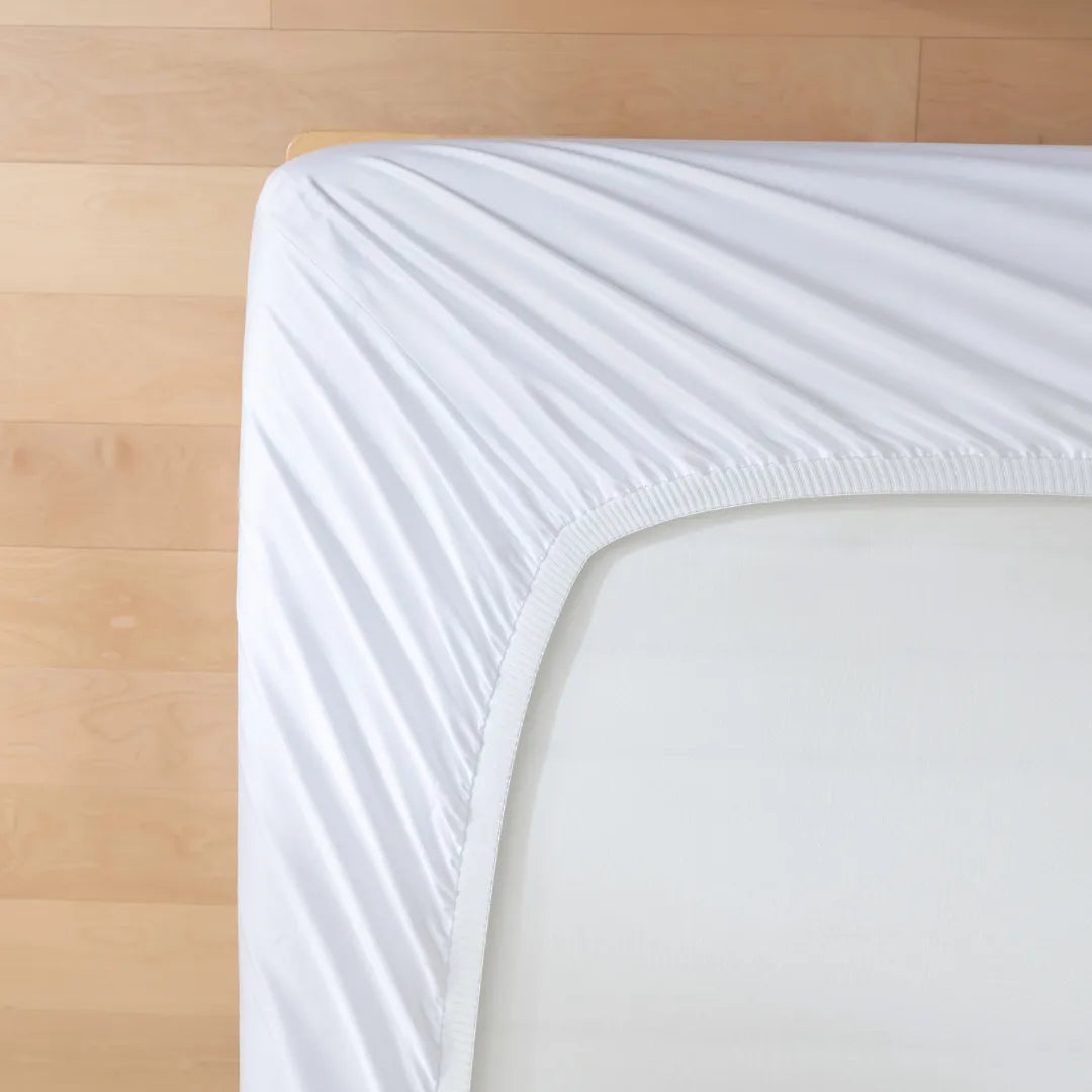 A neatly fitted white Linenly Luxe Sateen Sheet Set on a mattress with a smooth wooden floor in the background.